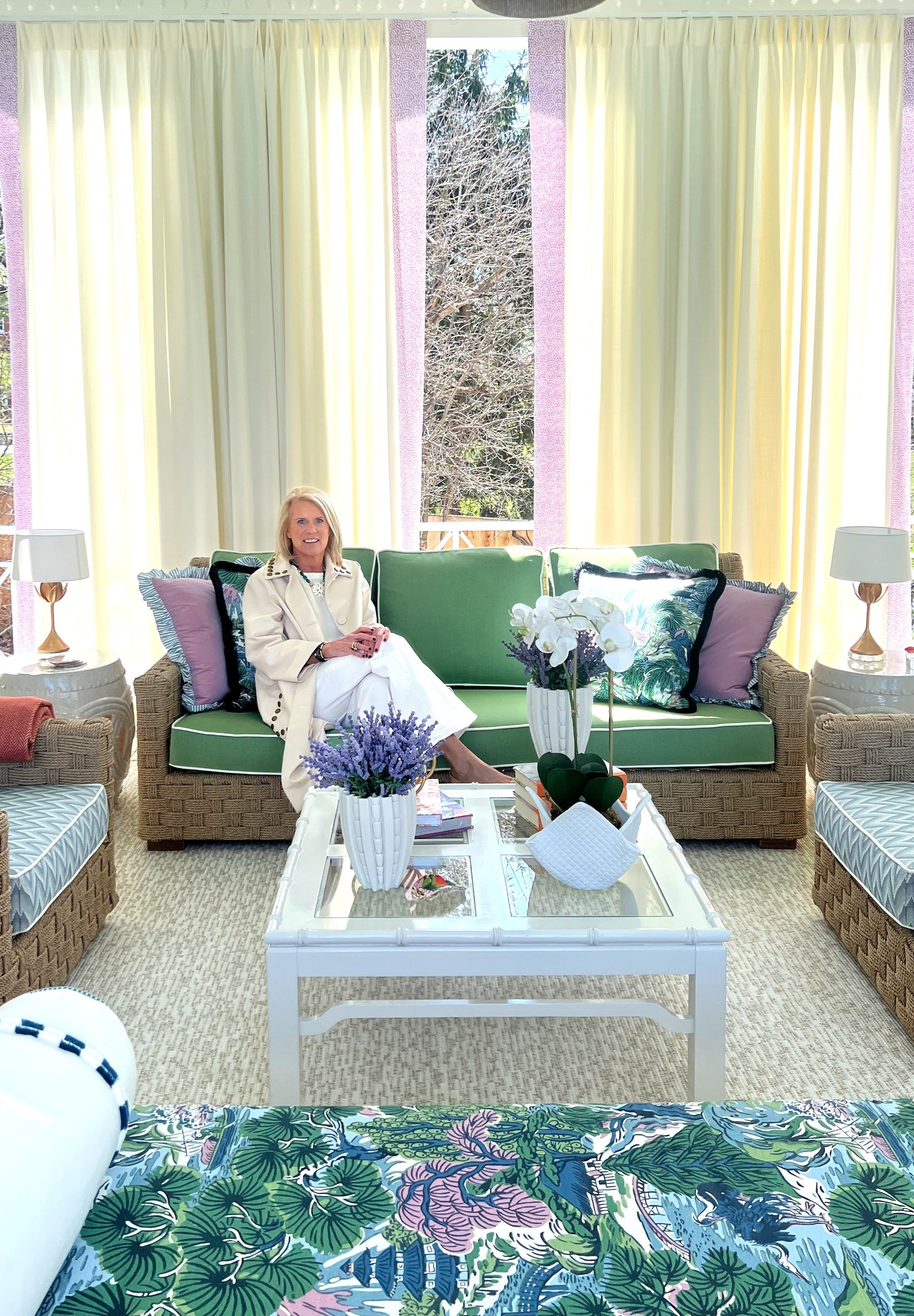 Woman sitting on a green sofa in a sunroom with large windows and decorative pillows.